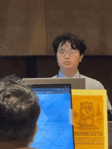 A person wearing glasses and a light blue shirt speaks from behind a laptop at a podium.