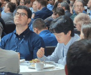 Two conference attendees sit at a round table during a large luncheon. One person in a navy shirt looks upward while another in a light blue shirt focuses on their meal. A crowd of other attendees fills the background.