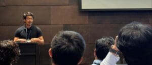 A speaker wearing glasses and a black shirt presents at a podium in front of an audience. The audience members’ heads are visible in the foreground while a large screen is positioned overhead.